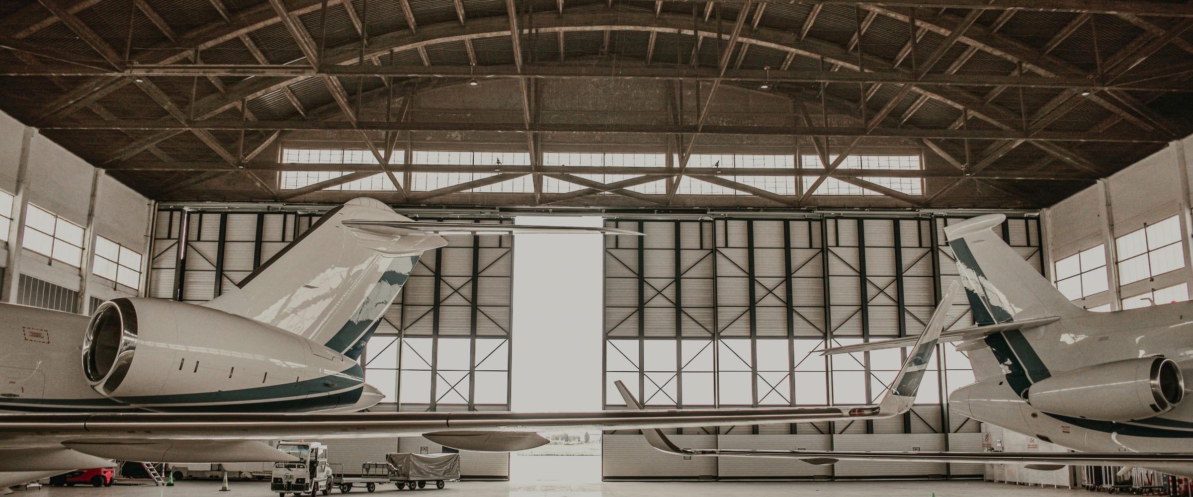 Two business jets nose-out in a hangar, tails almost touching, framed by the arched roof trusses and the daylight of the open hangar door between them.