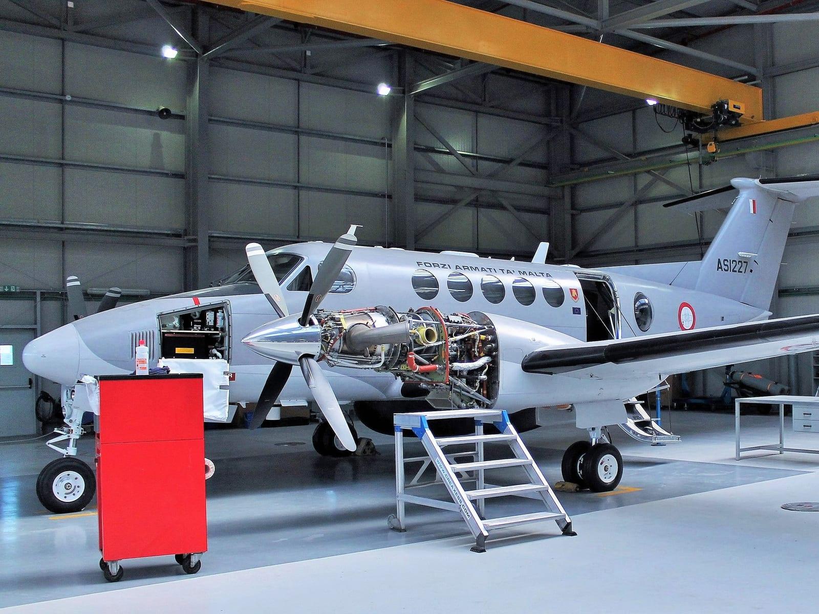 A King Air in a hangar mid-maintenance, one engine opened up and the nose avionics bay unpanelled.